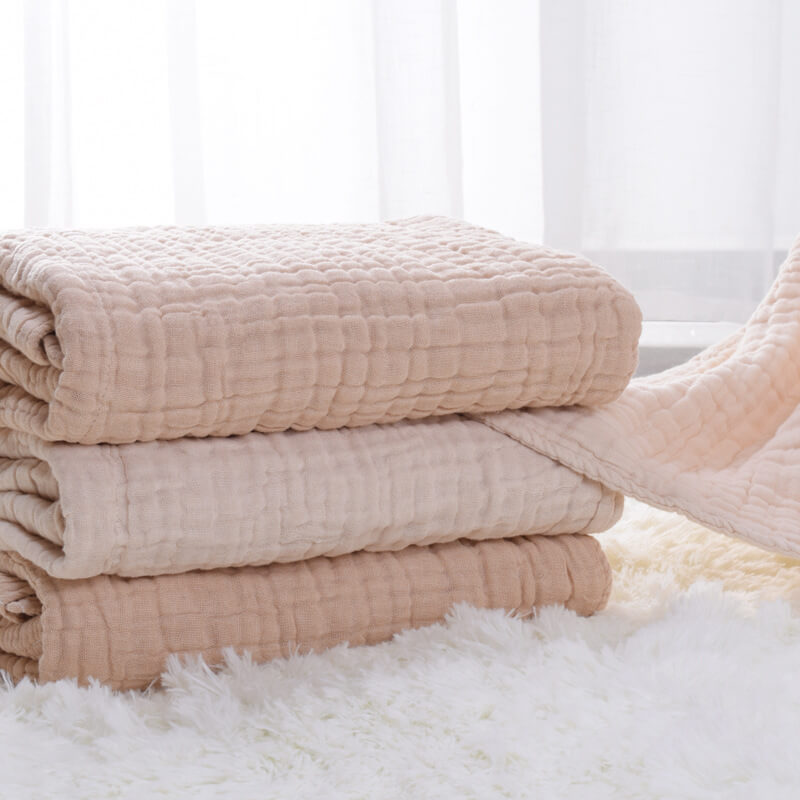 A stack of three neatly folded 6-layer organic colored cotton muslin baby blankets in natural beige, oatmeal, and ivory colors, highlighting the thickness.