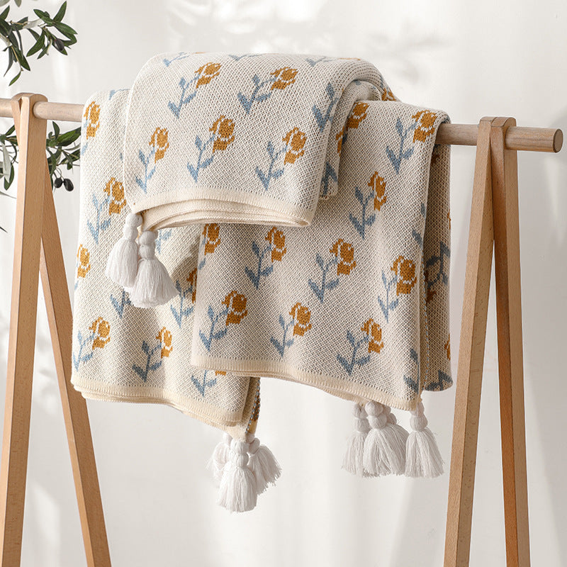 A floral acrylic knitted throw blanket with tassels, neatly folded and displayed on a wooden clothing rack in a well-lit room, showcasing the delicate floral pattern.