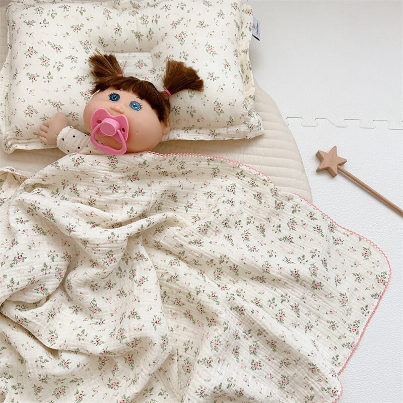 A muslin toddler blanket and pillow set with a Pomegranate Floral pattern, styled with a doll inside a crib. The soft, white blanket features a delicate print of red and grey floral motifs and has a pink scalloped edge.