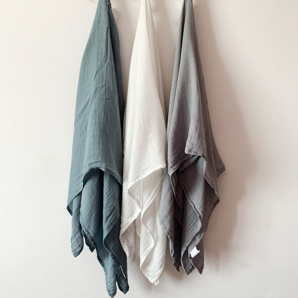 Three-pack of soft bamboo muslin swaddle blankets in cool tones, including teal, off-white, and slate gray, displayed against a clean white wall.