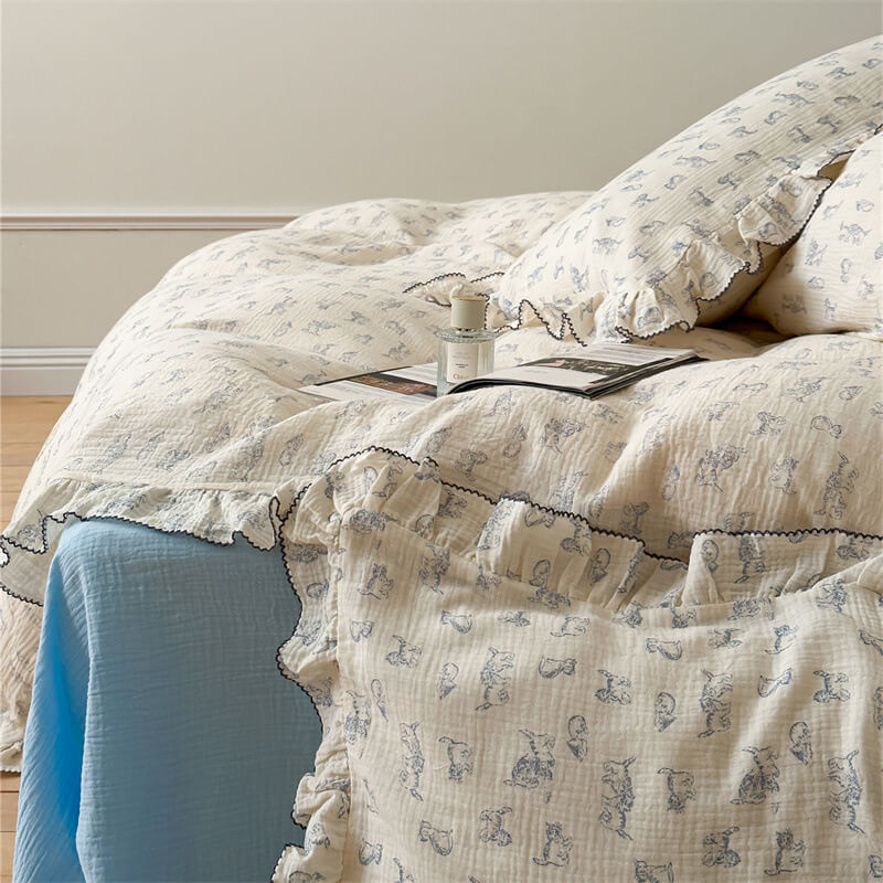 A close-up of a bed with a white cotton gauze duvet cover set, featuring a blue cat print and ruffled trim. A book and fragrance bottle rest on the duvet