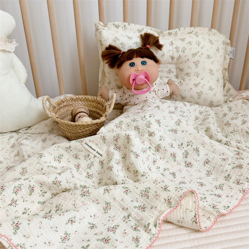A full view of the Pomegranate Floral muslin blanket and pillow set with a doll, tucked into a woven basket on a wooden surface.