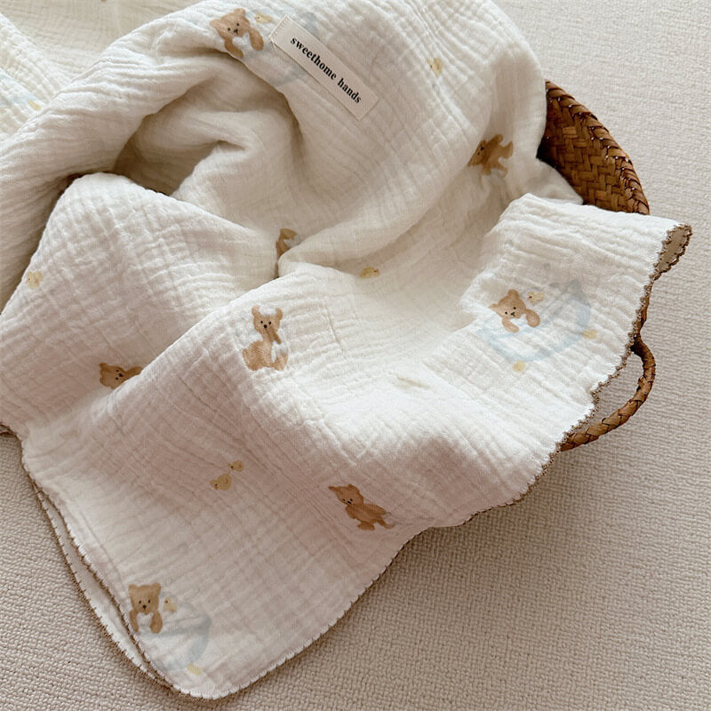 A white muslin toddler blanket with a teddy bear and soap bubble print, folded and placed inside a woven basket.