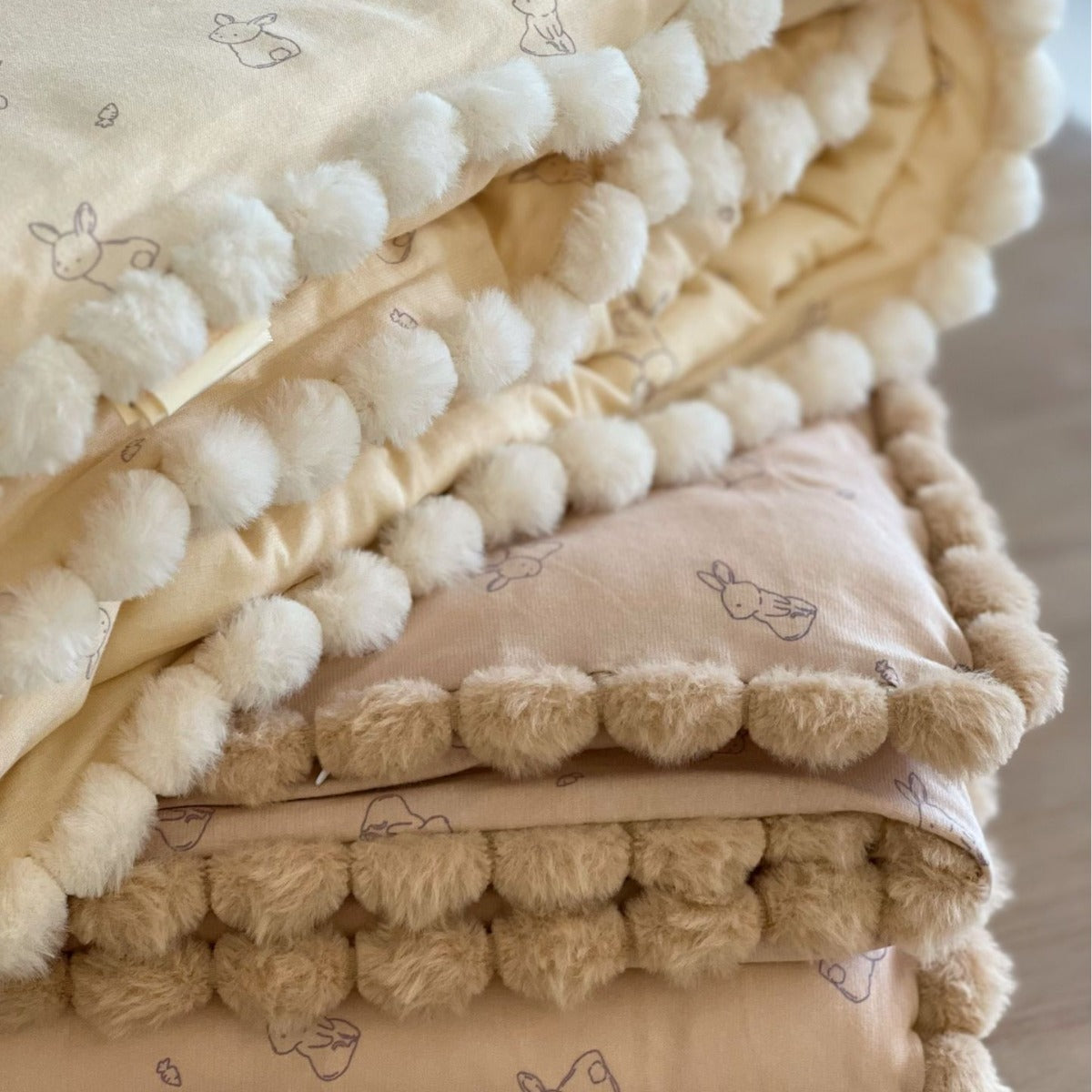 Stack of kids cotton comforter with pom-pom edges on a neutral background