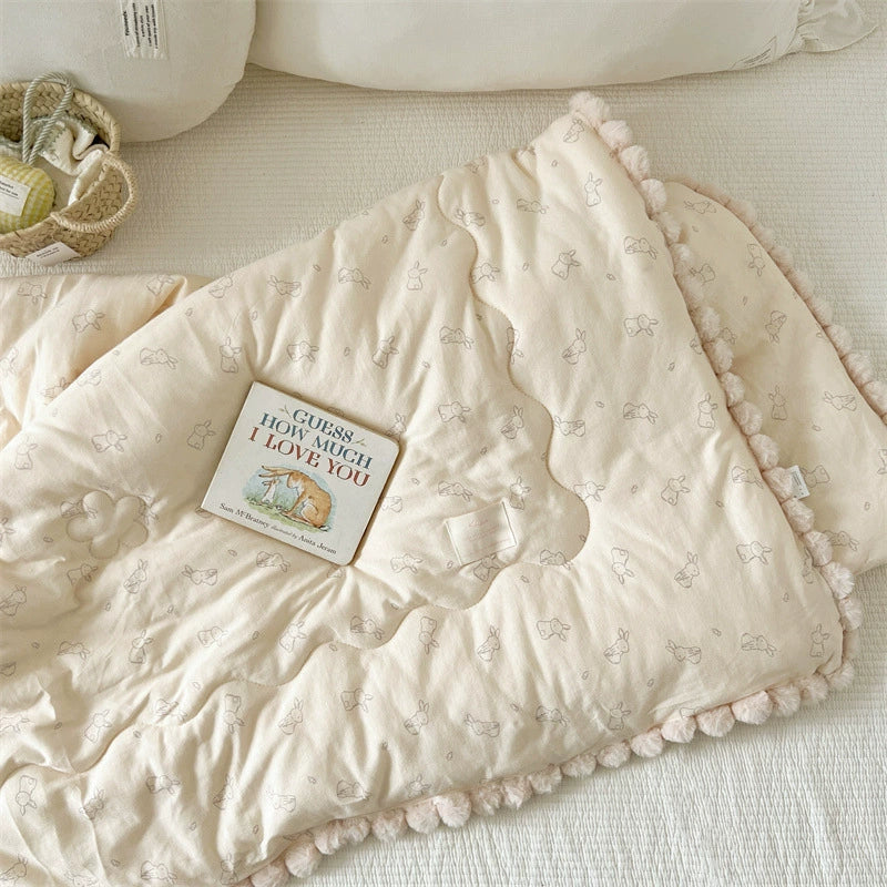 Beige cotton comforter with pom-pom edges and a book on a light surface.