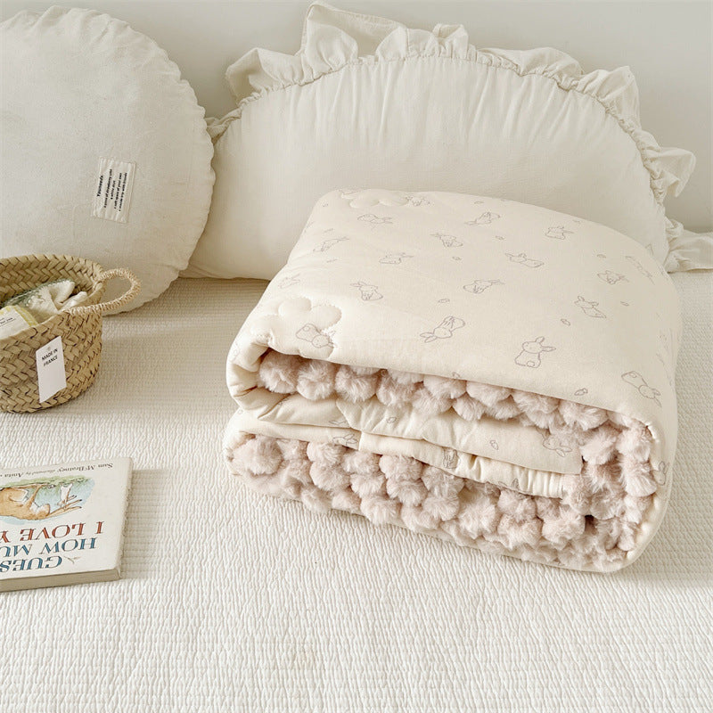Beige textured quilt with pom-pom trim on a light surface, next to a white pillow and small book.