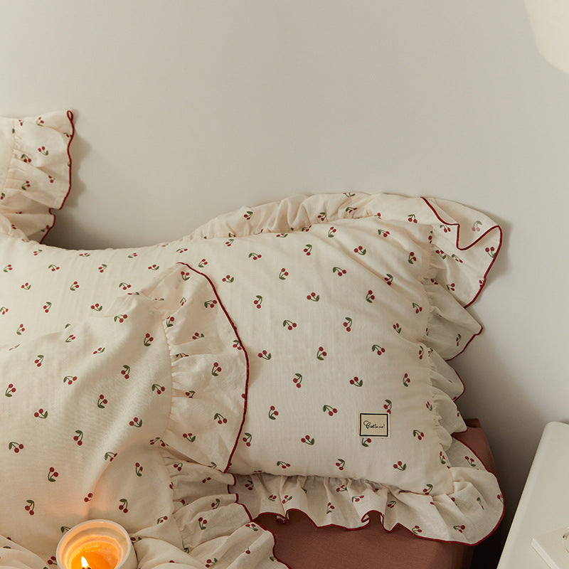 A close-up of the cherry-print cotton muslin pillowcase, featuring the delicate ruffle, red piping, and a brand label.
