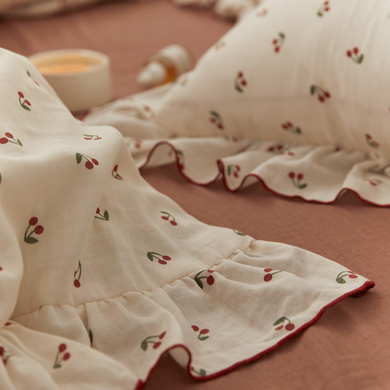 A cherry-print cotton muslin bedding set, including a ruffled pillow and comforter, folded and neatly stacked on a wooden surface.