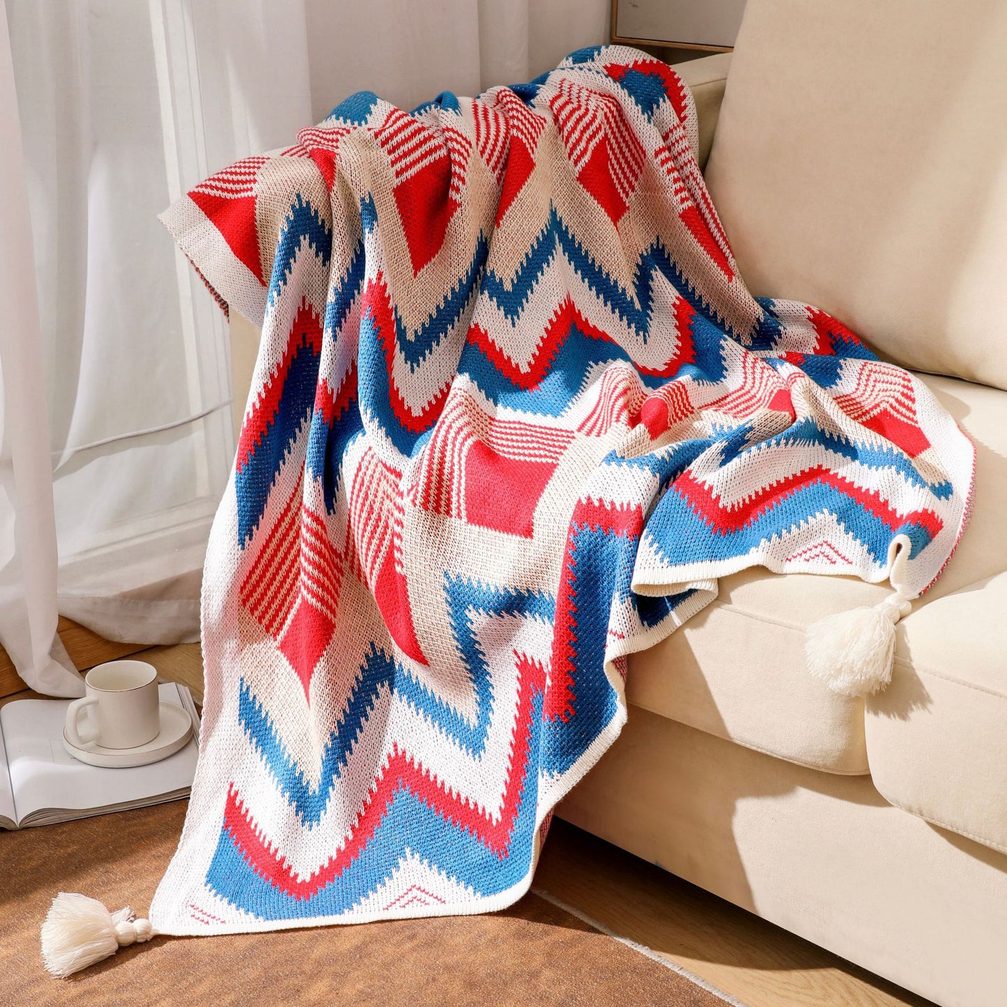 A vibrant red, white, and blue chevron knit boho throw blanket with tassels draped casually over the back of a light beige sofa in a sunlit living room.