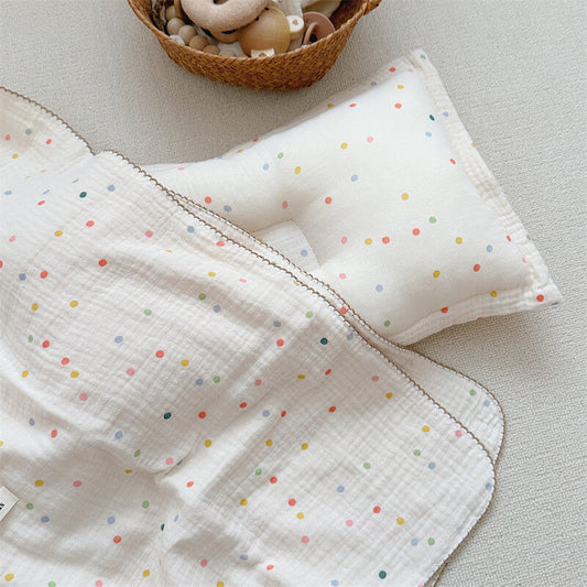 A top-down view of the Colorful Dot muslin toddler blanket and a matching pillow, showing the full set on a light-colored rug. A basket with toys is visible in the background.
