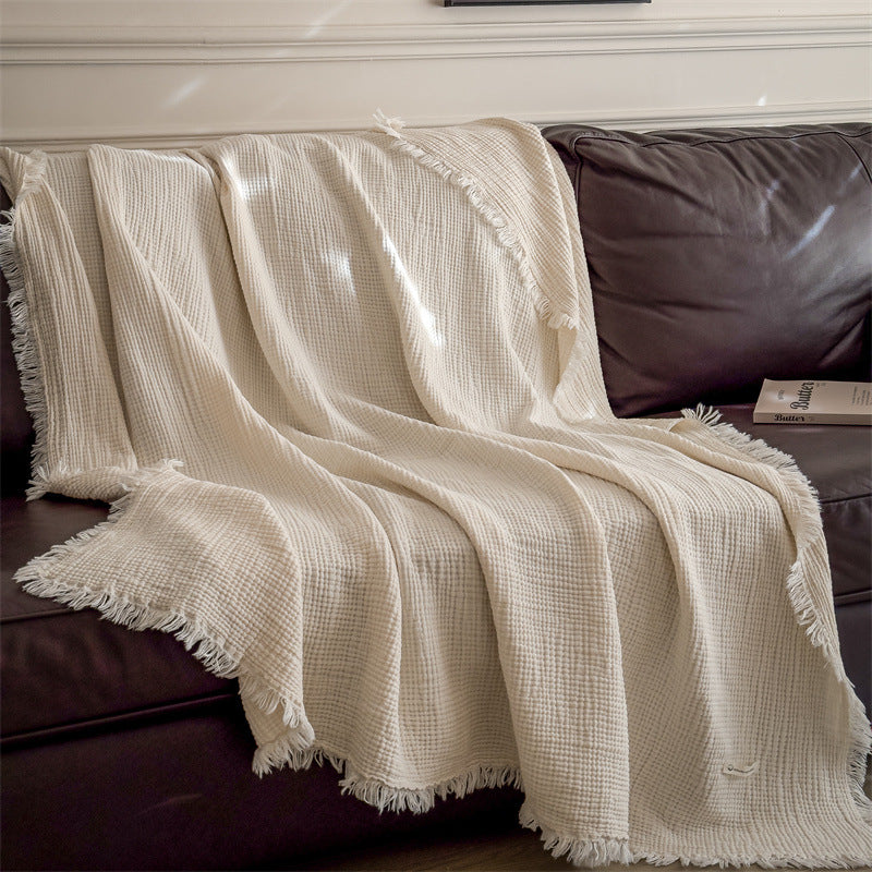 An ivory 4-layer cotton muslin throw blanket with fringe casually draped on a dark brown sofa, showcasing its soft and lightweight feel.