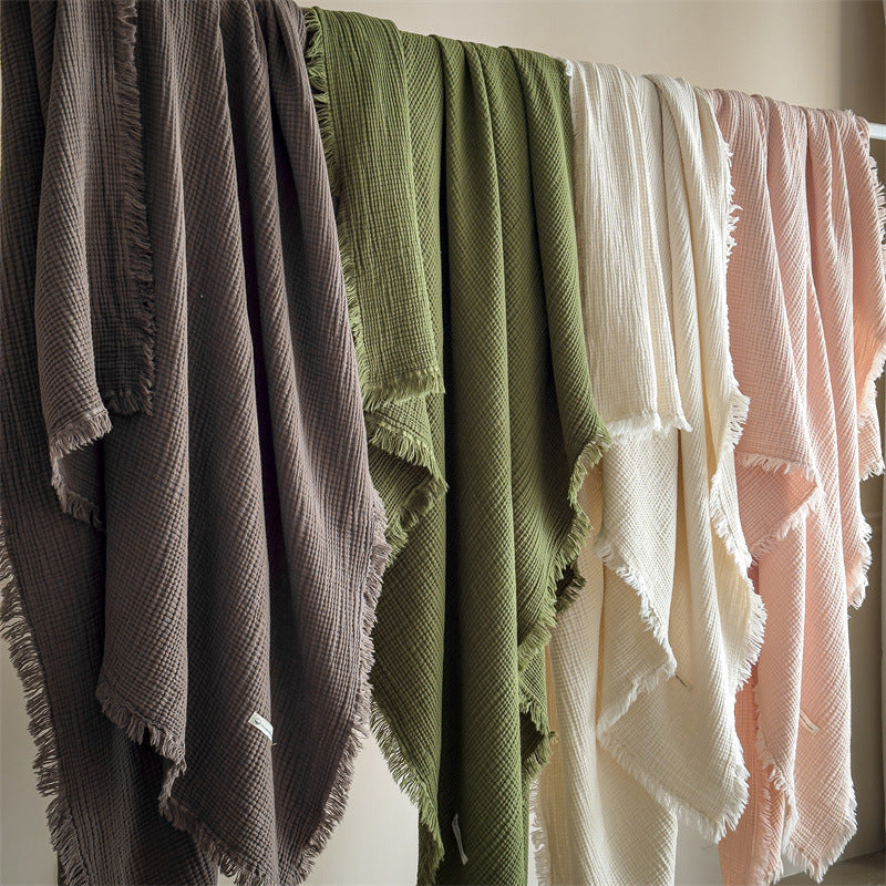 Multiple 4-layer cotton muslin throw blankets with fringe hanging on a rack, displaying different colors including brown, green, white, and pink.