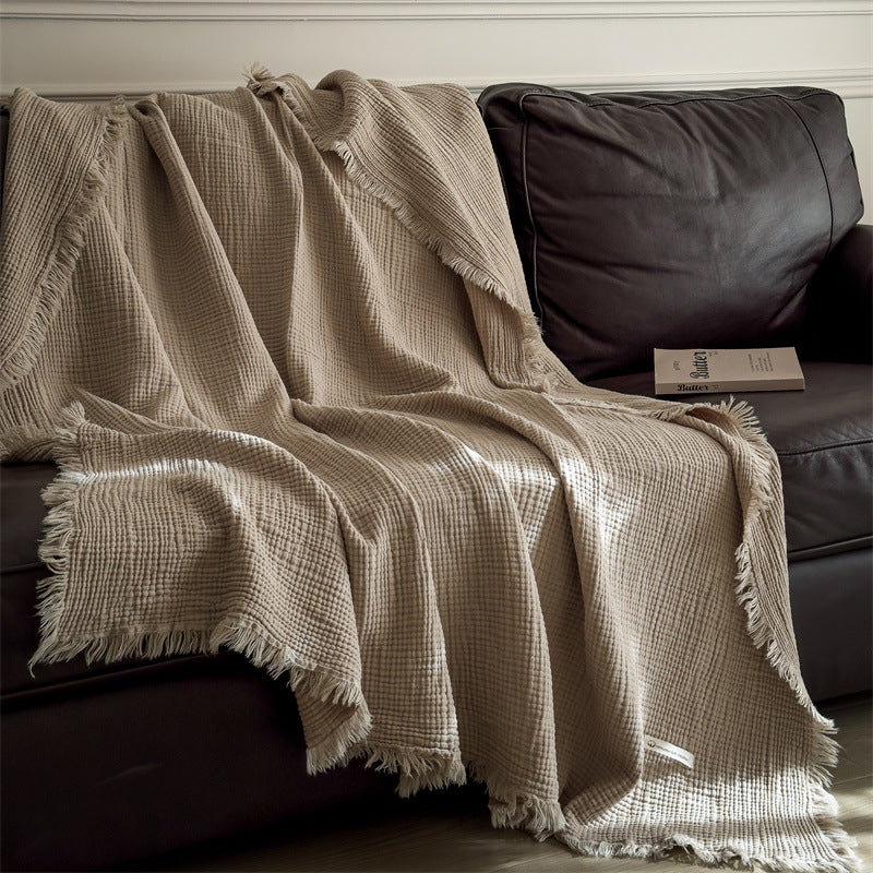 A beige 4-layer cotton muslin throw blanket with fringe draped neatly over a brown leather sofa.