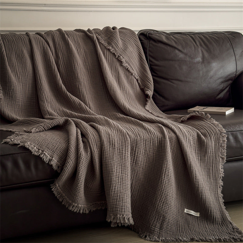 A dark brown 4-layer cotton muslin throw blanket with fringe casually draped over a brown leather sofa, adding warmth to the room.