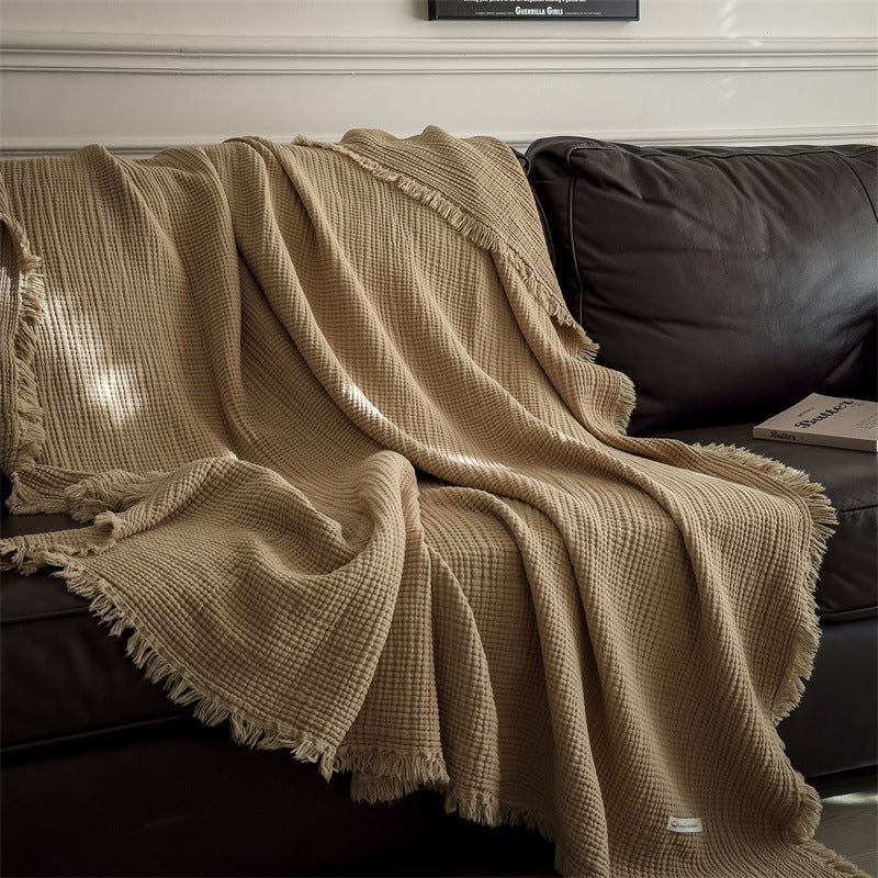 A light brown 4-layer cotton muslin throw blanket with fringe draped over a brown leather sofa, showing the blanket's soft folds and texture.