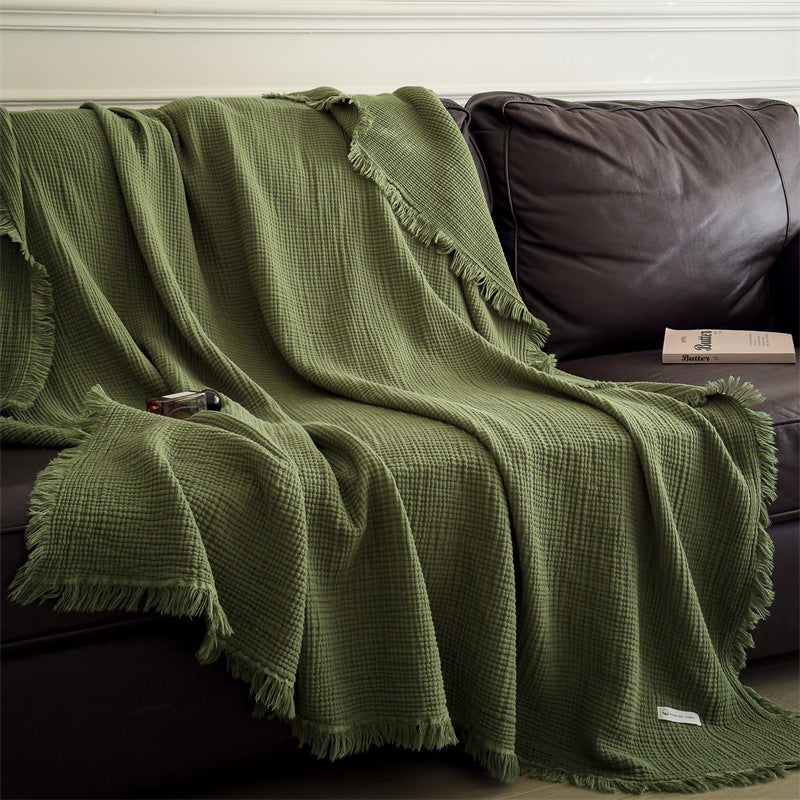 A stylish green 4-layer cotton muslin throw blanket with fringe draped over a brown leather sofa, providing a pop of color.