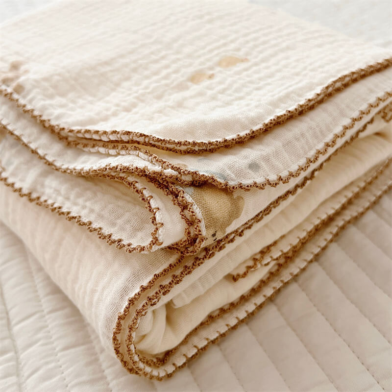 A close-up of a neatly folded cotton muslin toddler blanket with a brown scalloped edge, showing the texture of the fabric.
