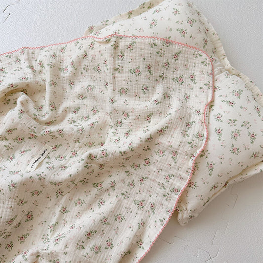 The folded Pomegranate Floral muslin blanket and pillow set, placed in a round, woven basket on a light-colored, textured rug.