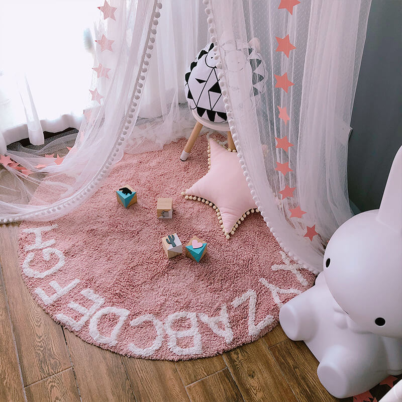 A pink-themed baby nursery with a hanging mesh crib mosquito net and a pink alphabet rug on the floor. The image shows the pink star pattern and the white pom-pom trim.