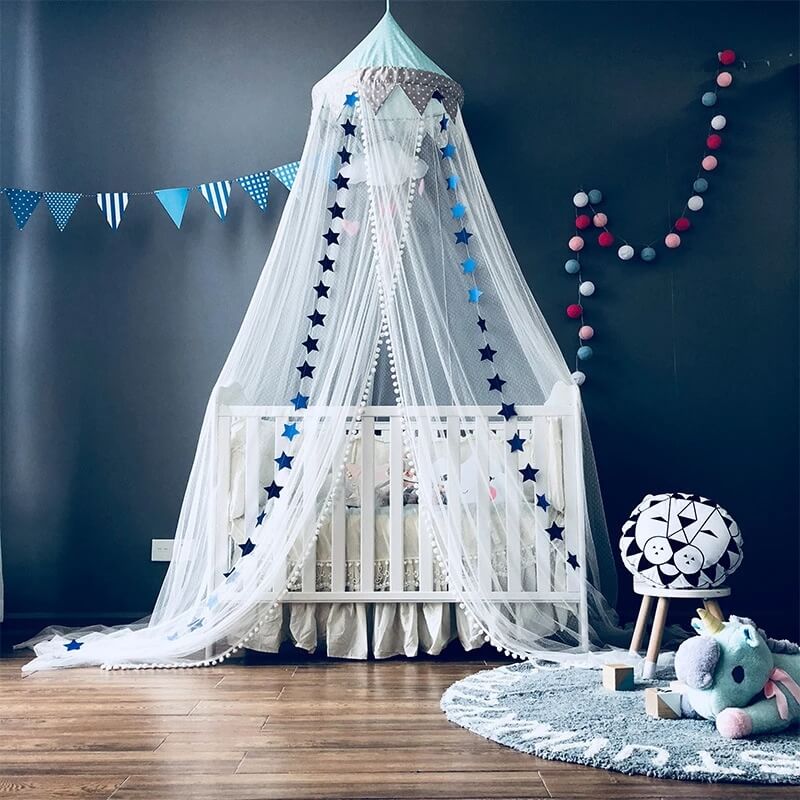 A hanging mesh crib mosquito net canopy with blue stars and white pom-pom trim, draped over a white crib in a nursery. The room features a dark gray wall, blue flag banner, and colorful pom-pom lights.