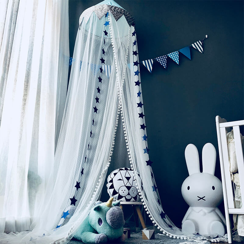A close-up view of a hanging mesh crib canopy with a blue star pattern and pom-pom trim, set in a baby's room with a Miffy lamp and lion-face stool.