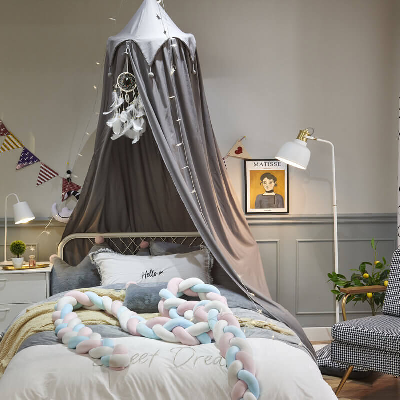 Gray kids bed canopy with falbala ruffles and pom-poms, over a bed with braided pillows and matching light gray decor.