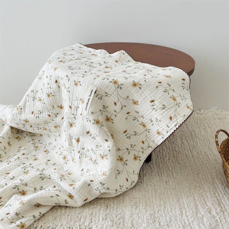 The yellow floral muslin baby blanket draped over a wooden table, showcasing the soft texture and pattern
