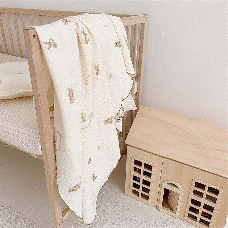 A teddy bear print muslin toddler blanket draped over the side of a light wooden crib, with a wooden dollhouse on the floor beside it.