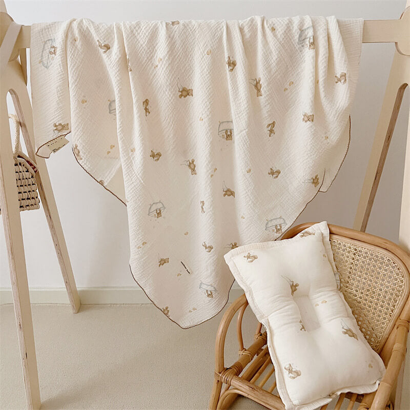 A muslin toddler blanket with a teddy bear and bubble bath print, draped over a wooden stand, with the matching pillow set on a rattan chair.