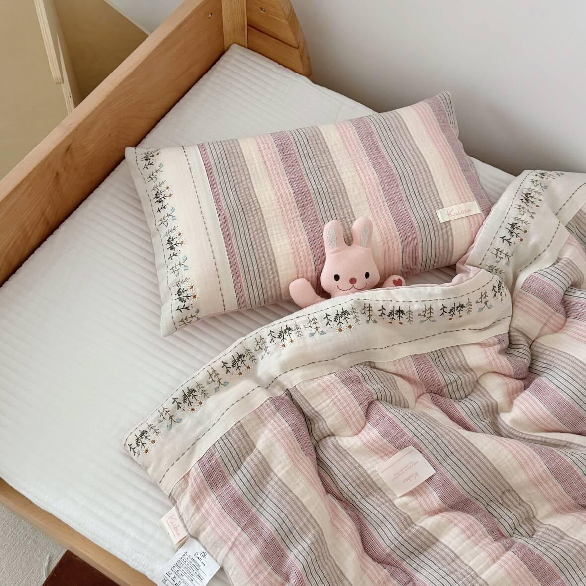 Pink striped muslin quilt set with delicate floral embroidery, paired with matching pillowcase on a wooden toddler bed