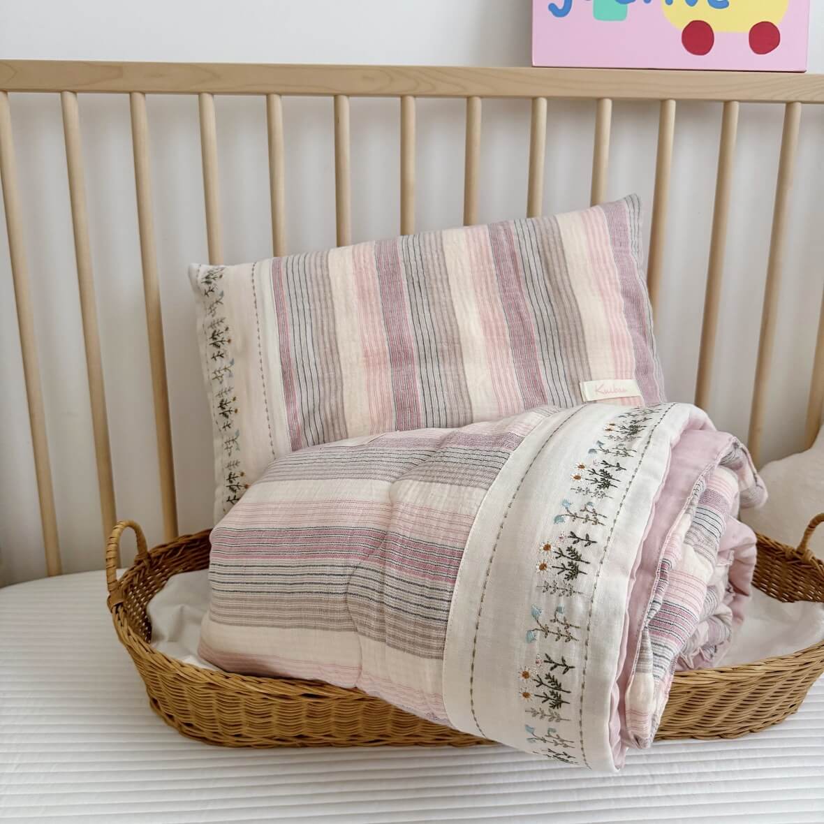 Striped pillowcase and quilt set in a wicker basket on a bed with a colorful poster in the background.