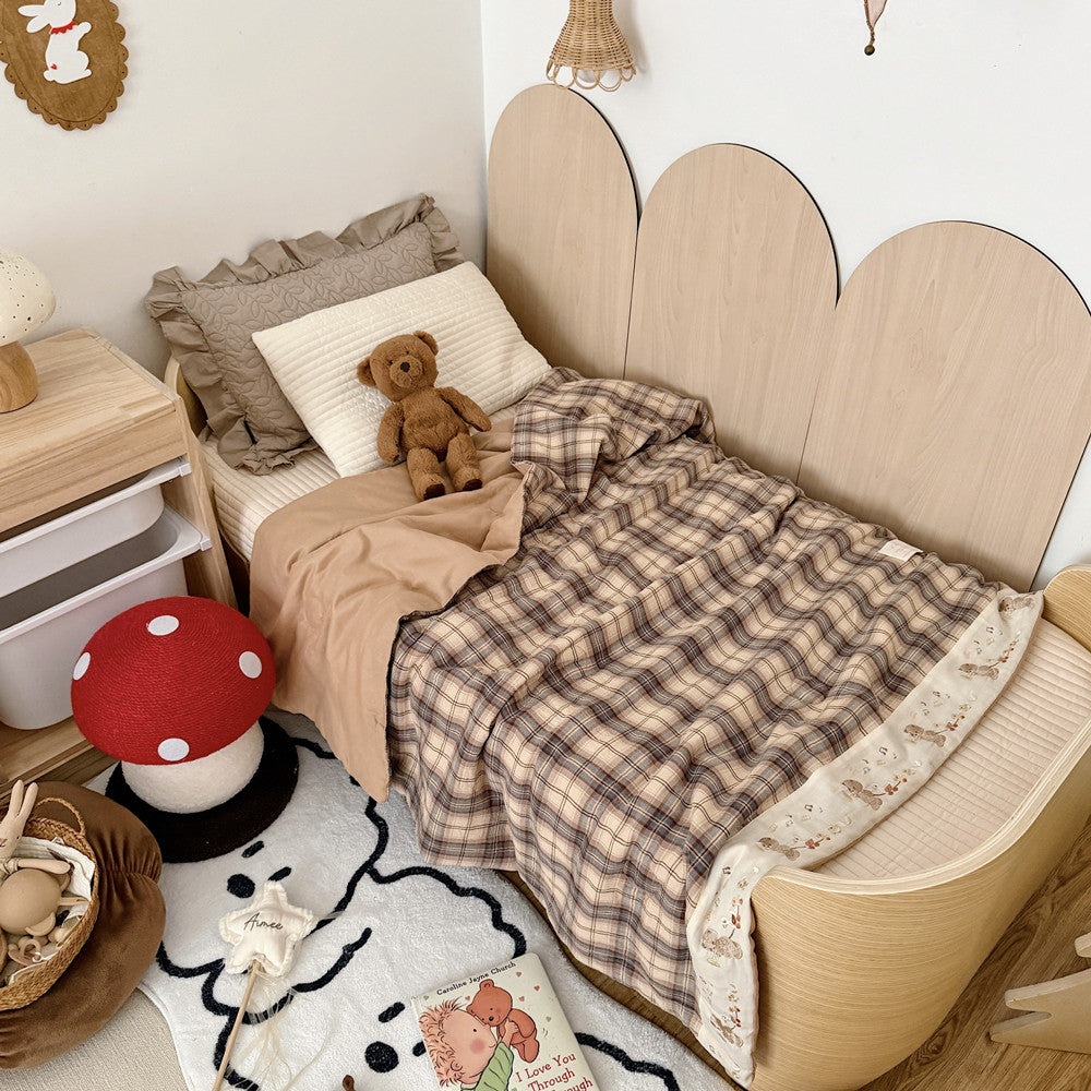 Plaid cotton muslin kids comforter with an embroidered cream border on a wooden bed beside a teddy bear.