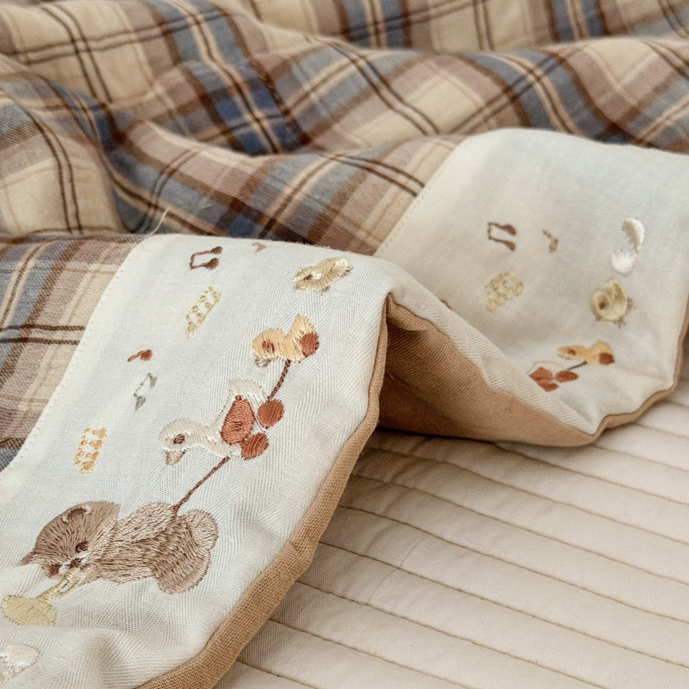 A close-up of a soft textile product, likely a blanket or quilt, showing a cream-colored front panel with embroidered woodland motifs including a fox, bird, and flowers, alongside a brown and beige plaid reverse side.