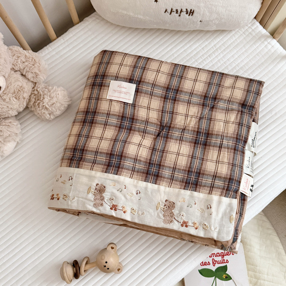 A folded plaid quilt or comforter with a beige, brown, and muted blue grid pattern, featuring a cream border with embroidered teddy bears and small floral details. It is displayed on a white quilted mattress in a nursery setting, surrounded by a plush teddy bear, wooden crib rails, and baby accessories.