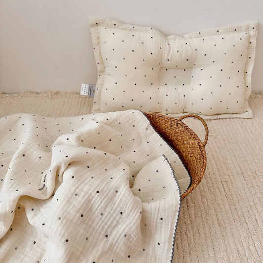 A close-up view of the Polka Dot muslin toddler blanket and pillow set, neatly folded in a woven basket on a textured blanket. The soft, neutral tones and subtle black and gray polka dots are visible.