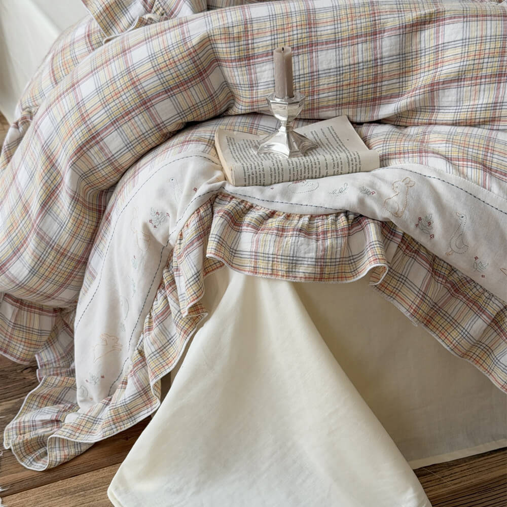 Detail of ruffled plaid duvet cover with embroidered bunny and duck trim, shown with a cream cotton flat sheet and decorative book on the bed