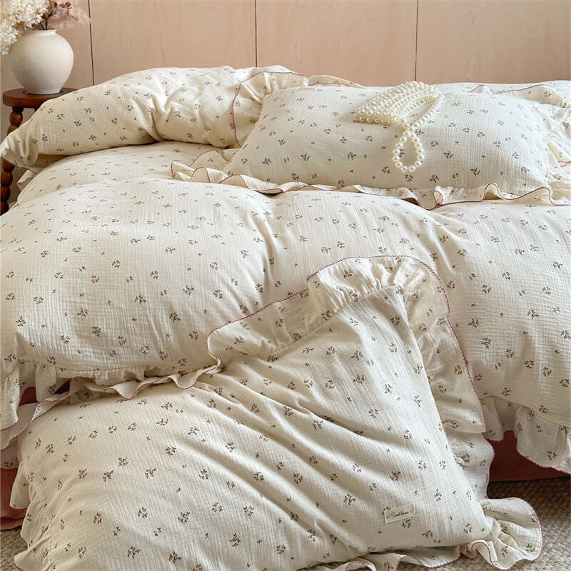 A close-up of a twin-size cotton muslin bedding set, showing the delicate floral print and the ruffled pillows with a pink scalloped trim.