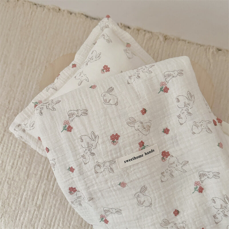 A warm-colored, cotton muslin toddler blanket and matching pillow set with a print of bunnies and flowers, shown folded on a light background.