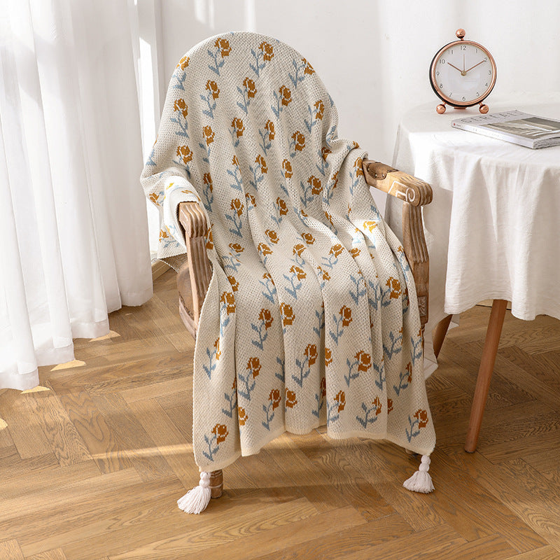 A floral acrylic knitted throw blanket with tassels draped over a vintage wooden chair next to a small table, creating a cozy and decorative accent.