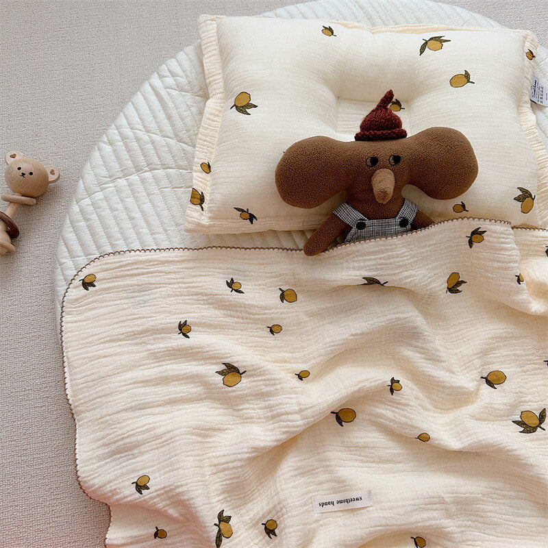 A cozy baby bedspread with a pillow set, showcasing a lemon print, neatly laid out on a white mat with a brown plush toy tucked under the blanket.