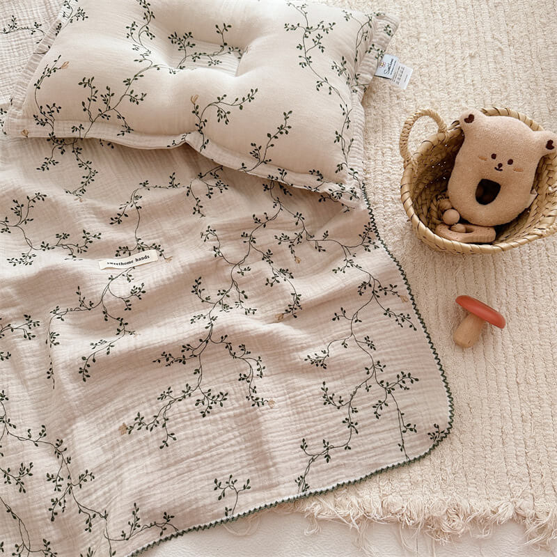 A flat-lay photo of the Botanical muslin blanket and pillow, showing their placement on a light-colored rug with a small basket and toys nearby.