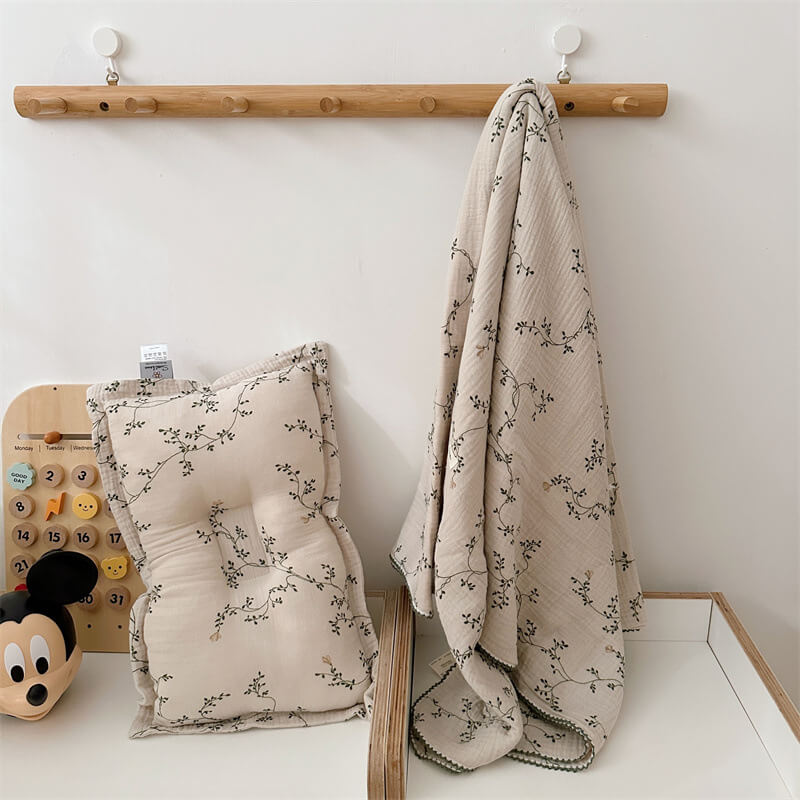 The Botanical muslin toddler blanket and pillow set, 47"x51", displayed on a changing table with a matching wall-mounted hook rack.