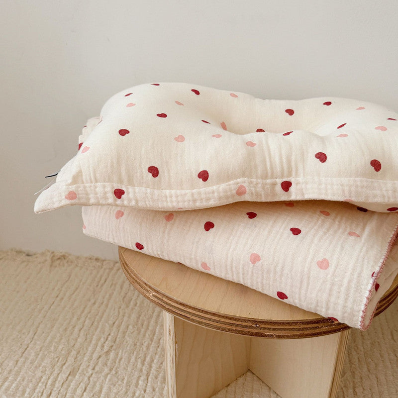 A side view of the Pink and red Heart muslin blanket and pillow set, stacked on a small wooden stool, showcasing the layered design.