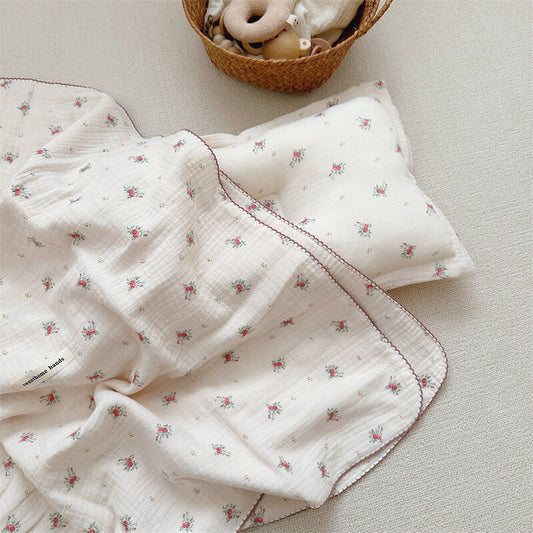 A baby girl floral receiving blanket from the Tiny Rose collection, neatly folded next to its matching pillow with a woven basket of toys in the background.