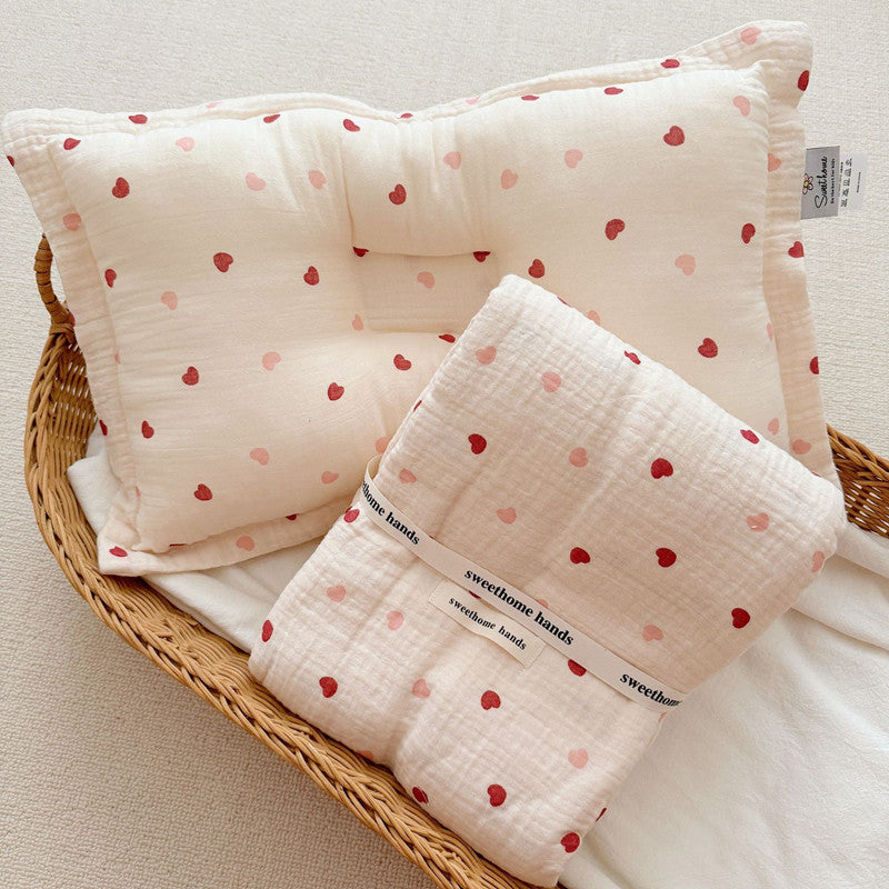 The Pink Heart muslin blanket and pillow set, neatly folded in a woven basket, highlighting the light pink and red heart pattern.