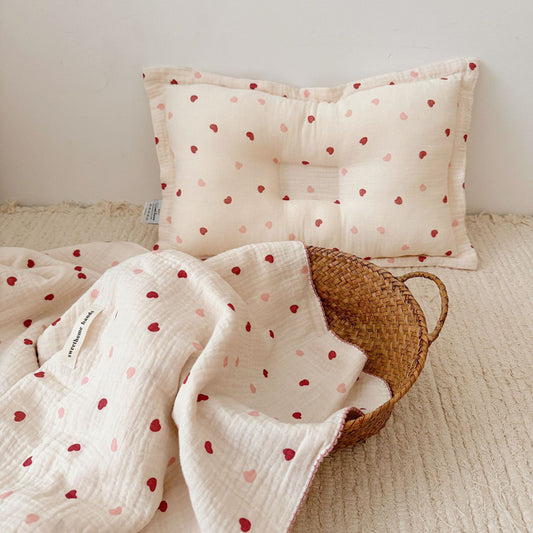 A Pink and red Heart muslin toddler blanket and pillow set, 47"x51", in a crib with a plush toy and a star rattle. This image shows a complete set for a baby's nursery.