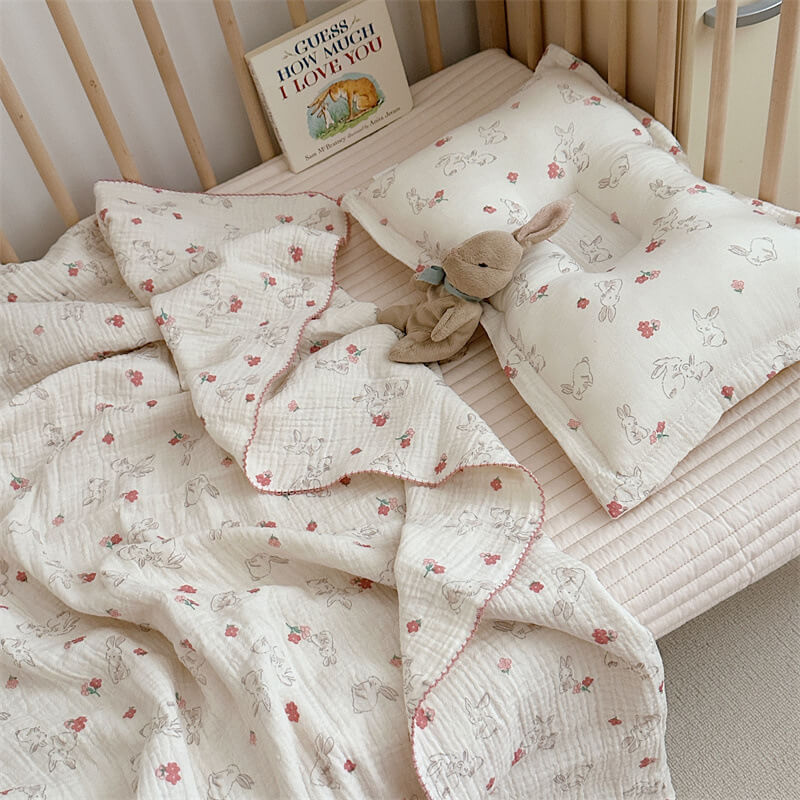 An overhead view of a baby girl's crib featuring a white cotton muslin blanket and pillow set with a bunny and floral pattern, along with a stuffed bunny toy