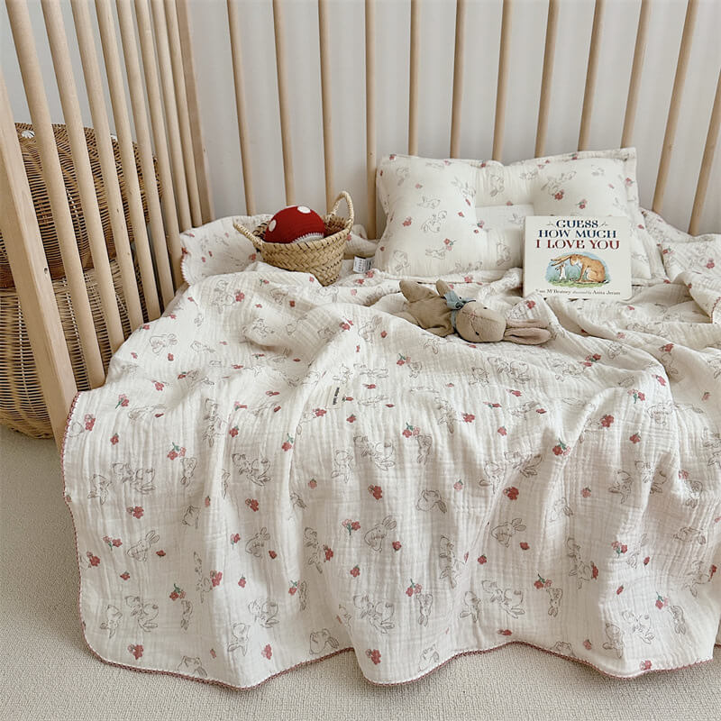 A baby girl's blanket and pillow set with a bunny and floral pattern, laid out inside a wooden crib with a plush toy and a children's book.