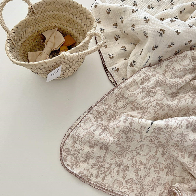 A Dreamwoods floral cotton muslin baby blanket with a pink scalloped edge, alongside a floral blanket and a small wicker basket, perfect for a baby girl.
