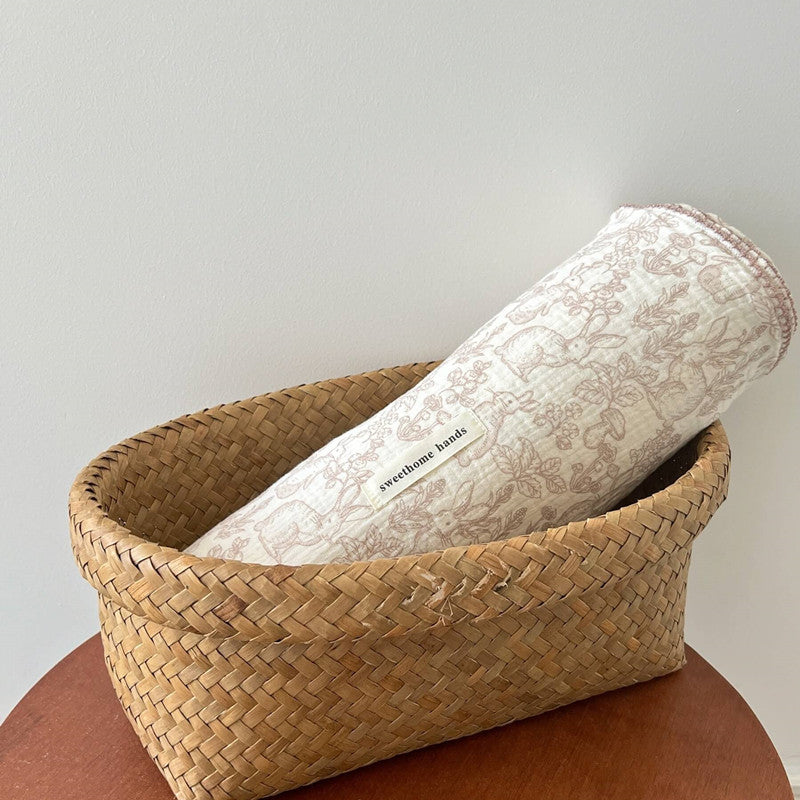 A rolled-up Dreamwoods floral muslin baby receiving blanket tucked into a natural wicker basket, ideal for infants.